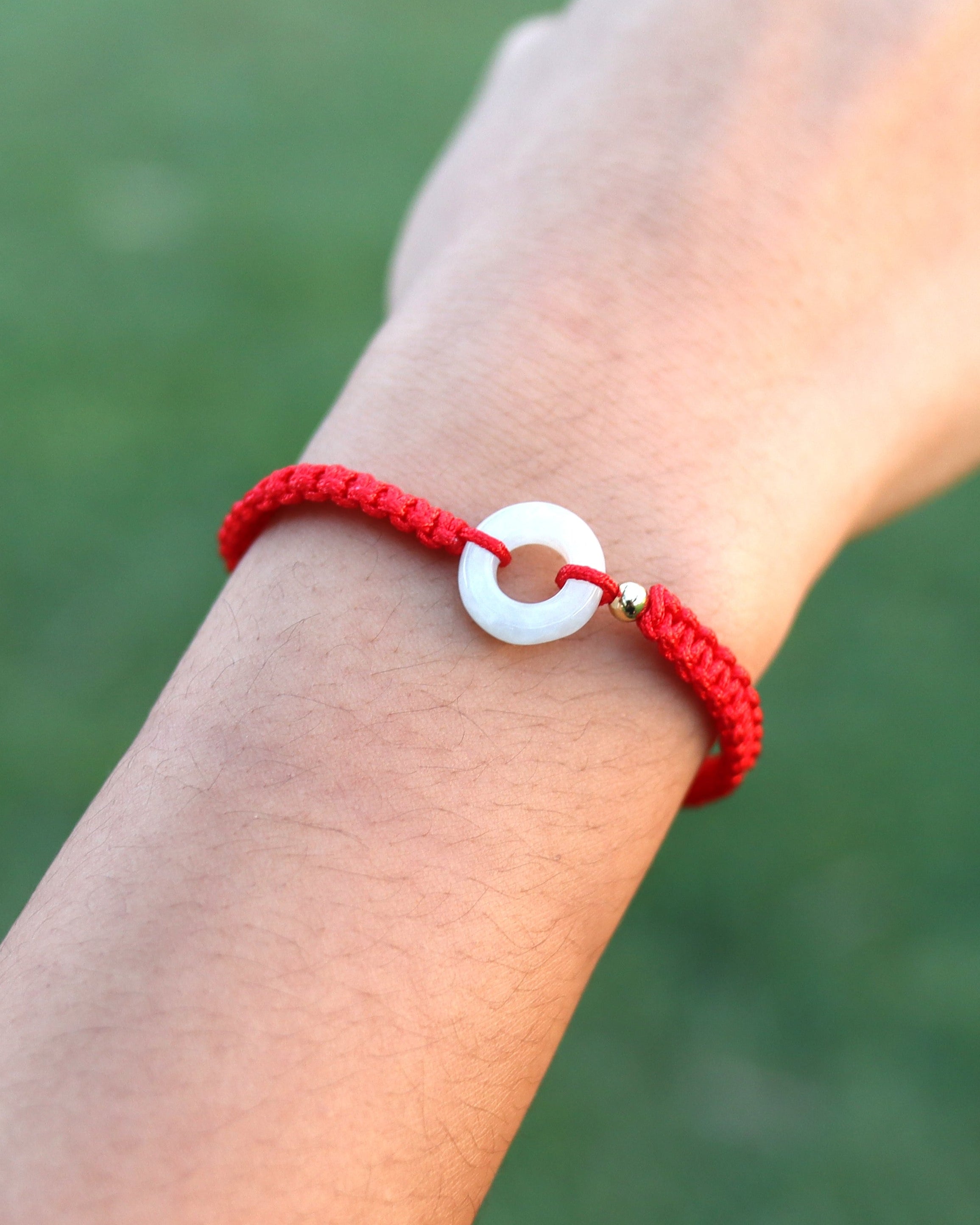 A red braided cord bracelet with a white circular charm, worn on a person's wrist against a blurred green outdoor background.