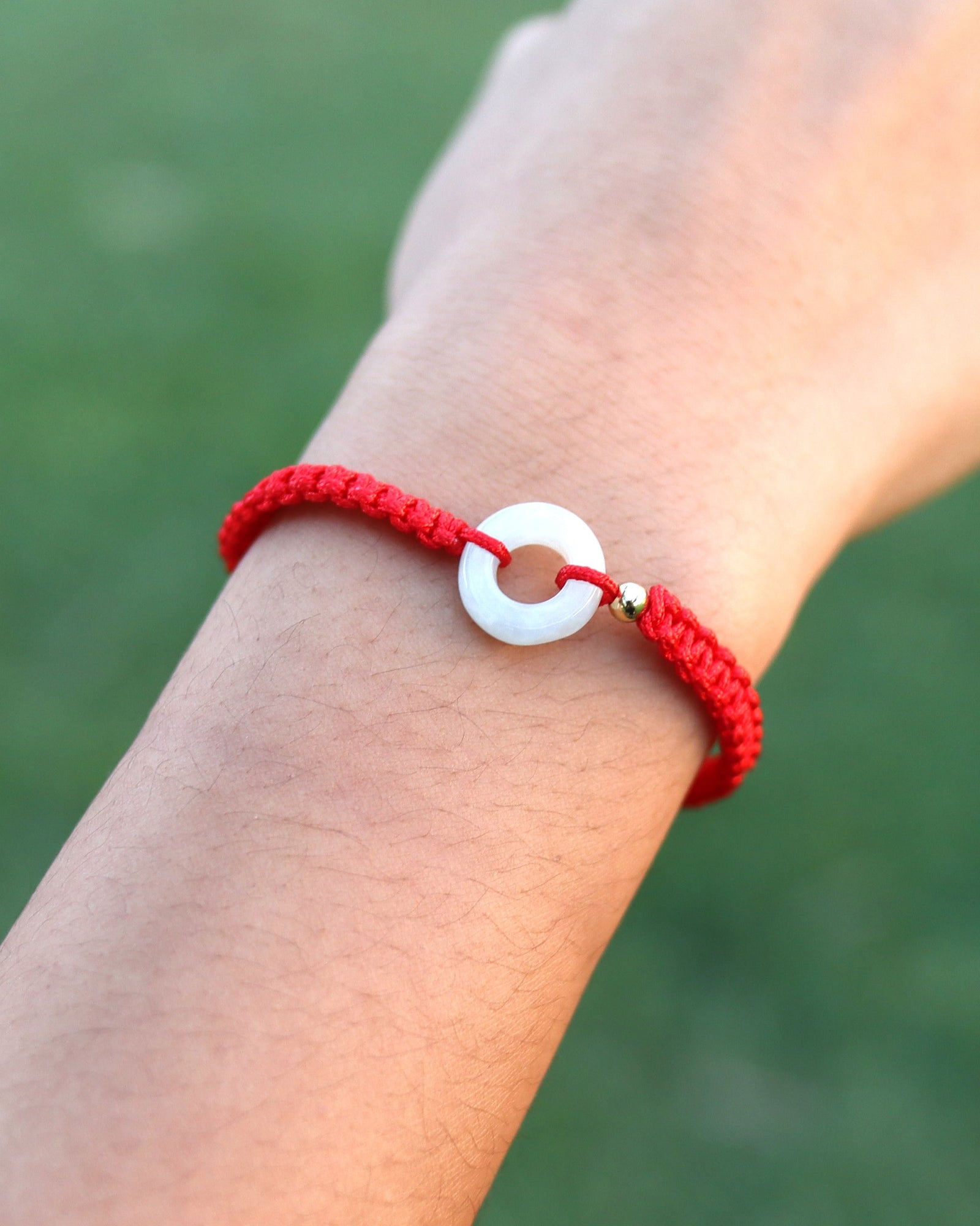A red braided cord bracelet with a white circular charm, worn on a person's wrist against a blurred green outdoor background.